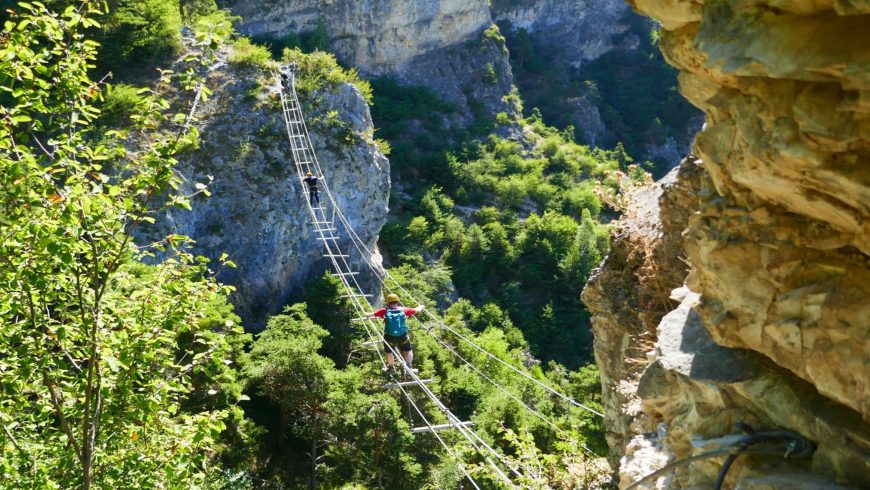 Un étroit pont suspendu traverse une gorge rocheuse, entourée d'une végétation luxuriante et de falaises spectaculaires.